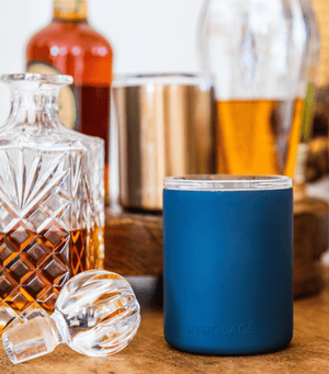A blue glass lined whiskey low ball glass with a stainless steel exterior, displayed alongside a decanter of whiskey and ice cubes.
